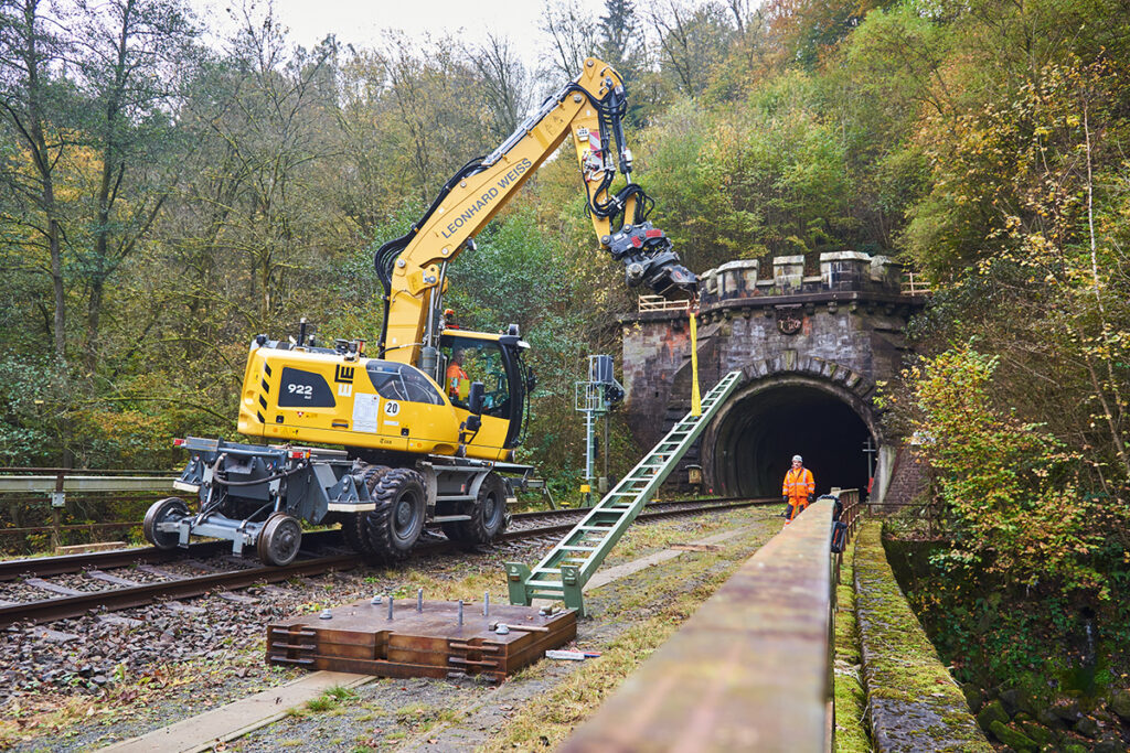 Ein Bagger steht auf einem Gleis vor einem Tunnel und hebt einen Oberleitungsmast an.