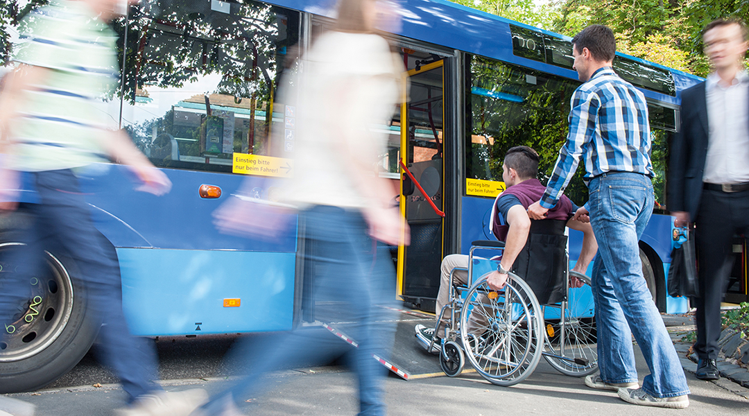 Eine Personen schiebt eine andere Person in einem Rollstuhl in einen Bus.