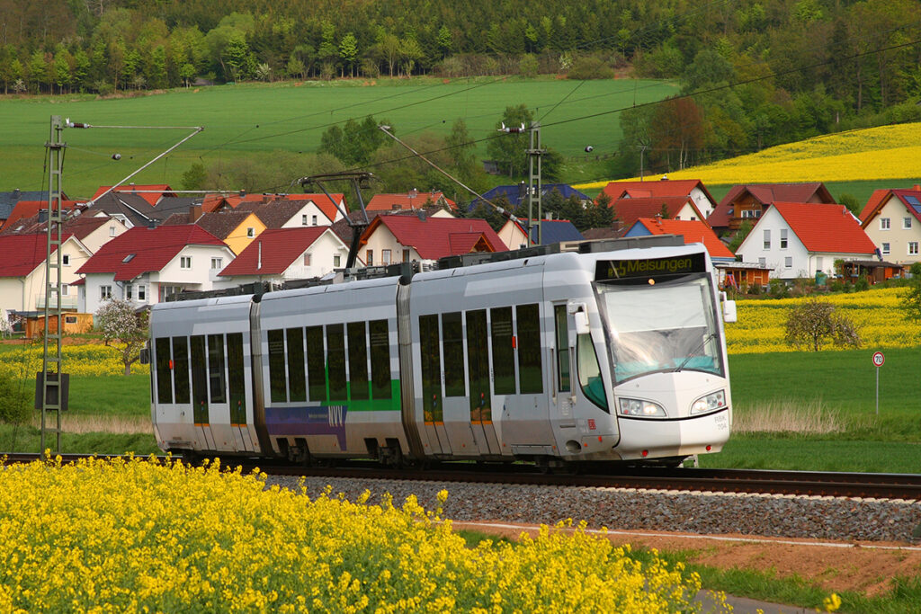 Eine Stadtbahn fährt an Feldern und Häusern entlang.