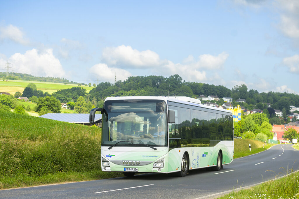Ein Bus fährt auf einer Straße vor Wiesen und einem See.