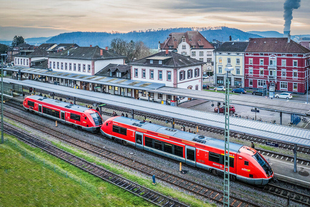 Ein Zug steht in einem Bahnhof, hinter dem Empfangsgebäude stehen weitere Häuser.