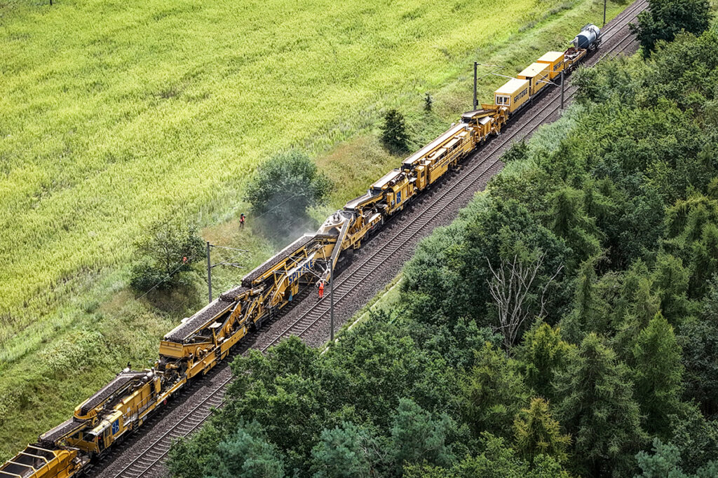 Ein Bauzug steht auf Gleisen zwischen Wiesen und Wald.