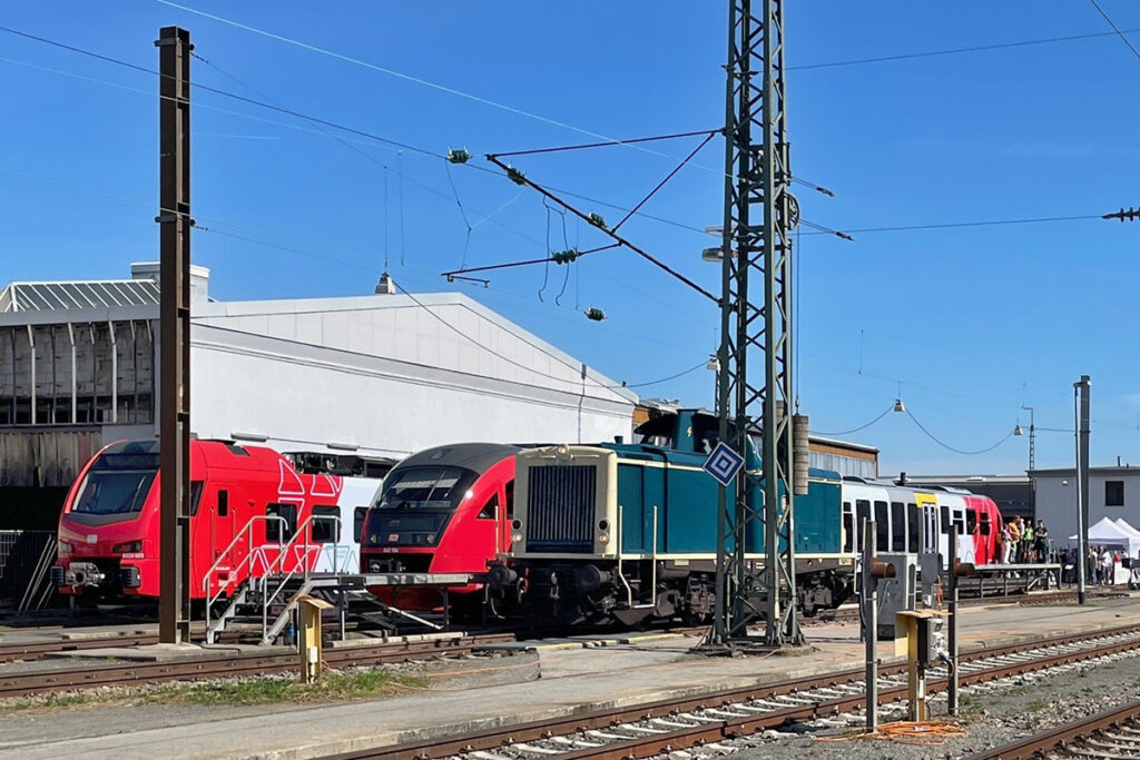 Eine Diesellok und Triebzüge stehen vor einer Halle.