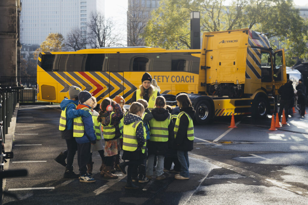 Grundschulkinder stehen mit ihrer Lehrerin vor einem LKW und vor einem Bus