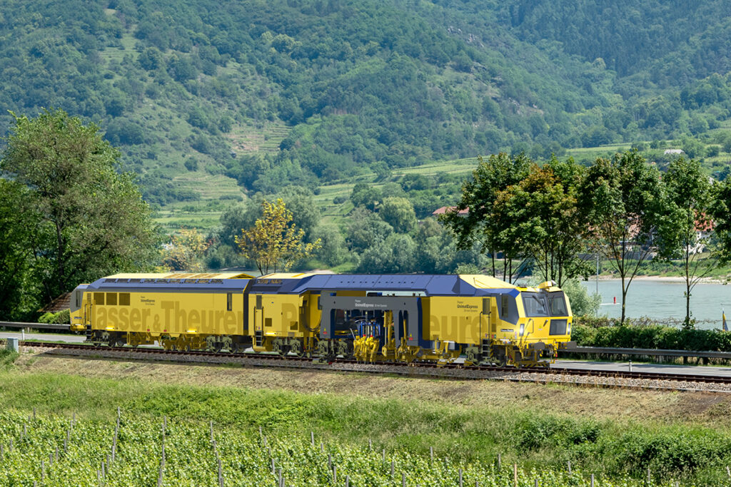 Simulation einer Stopfmaschine auf einem Gleis vor Bäumen.