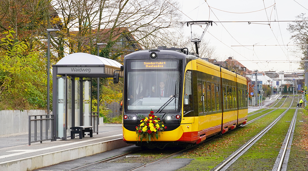 Eine Tram hält an einer Haltestelle vor Bäumen.