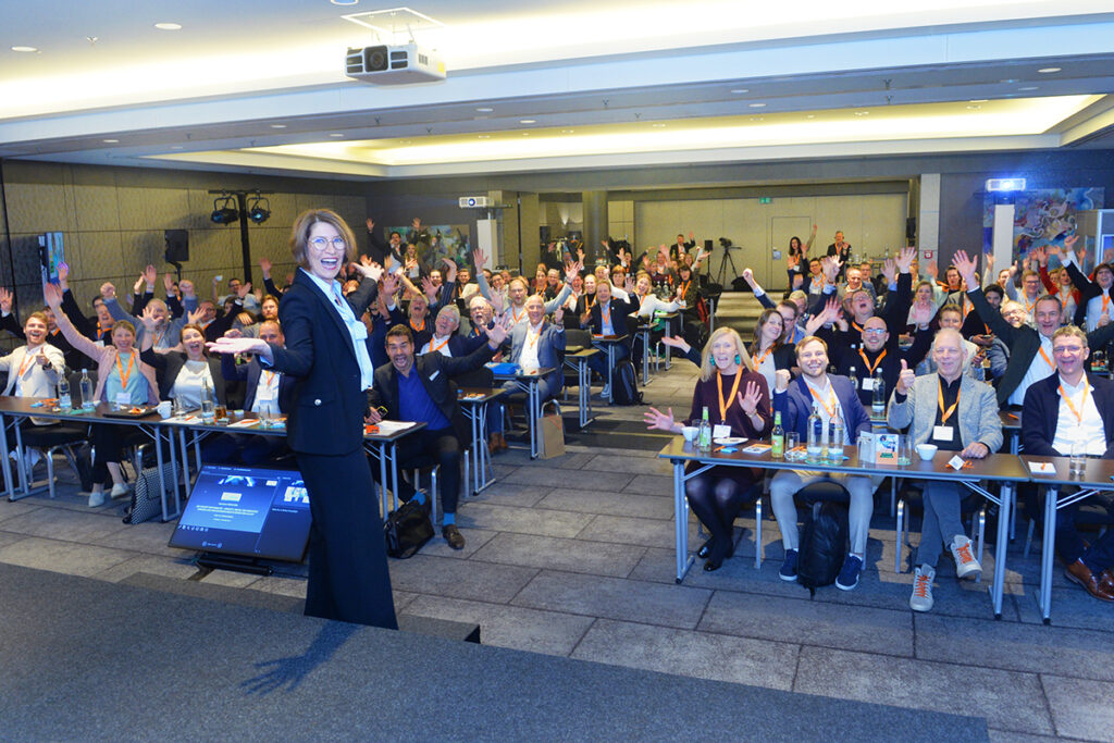 Viele Menschen, die an Tischen sitzen, winken in die Kamera.