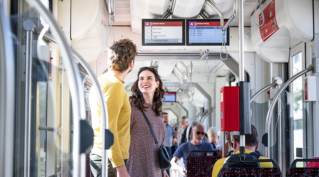 Zwei Personen stehen in einer Straßenbahn unter einer Anzeige.