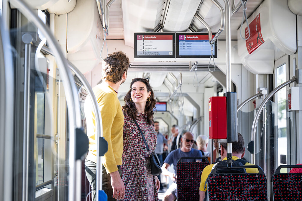 Zwei Personen stehen in einer Straßenbahn unter einer Anzeige.