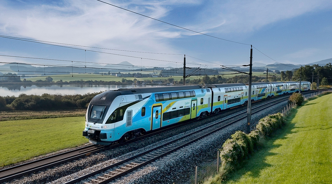 Ein Zug fährt auf einer zweigleisigen Strecke an Wiesen und einem See vorbei.