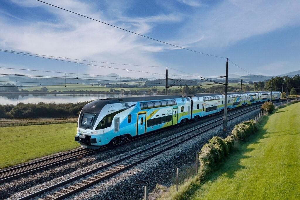 Ein Zug fährt auf einer zweigleisigen Strecke an Wiesen und einem See vorbei.