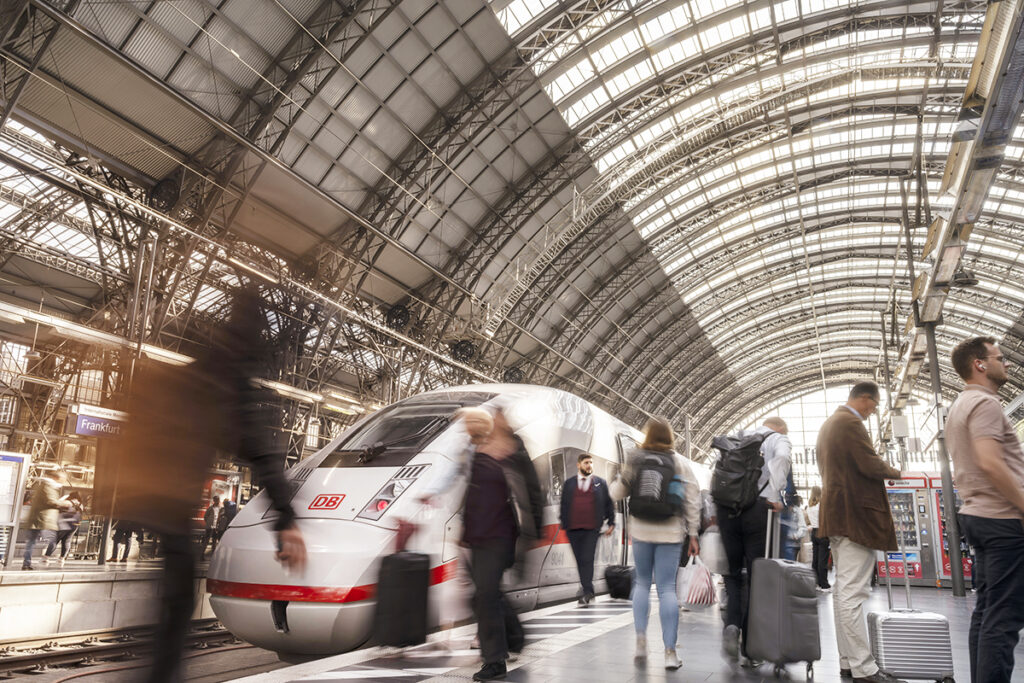 Ein ICE hält an einem Bahnsteig in einer Bahnhofshalle, auf dem Bahnsteig stehen Reisende.