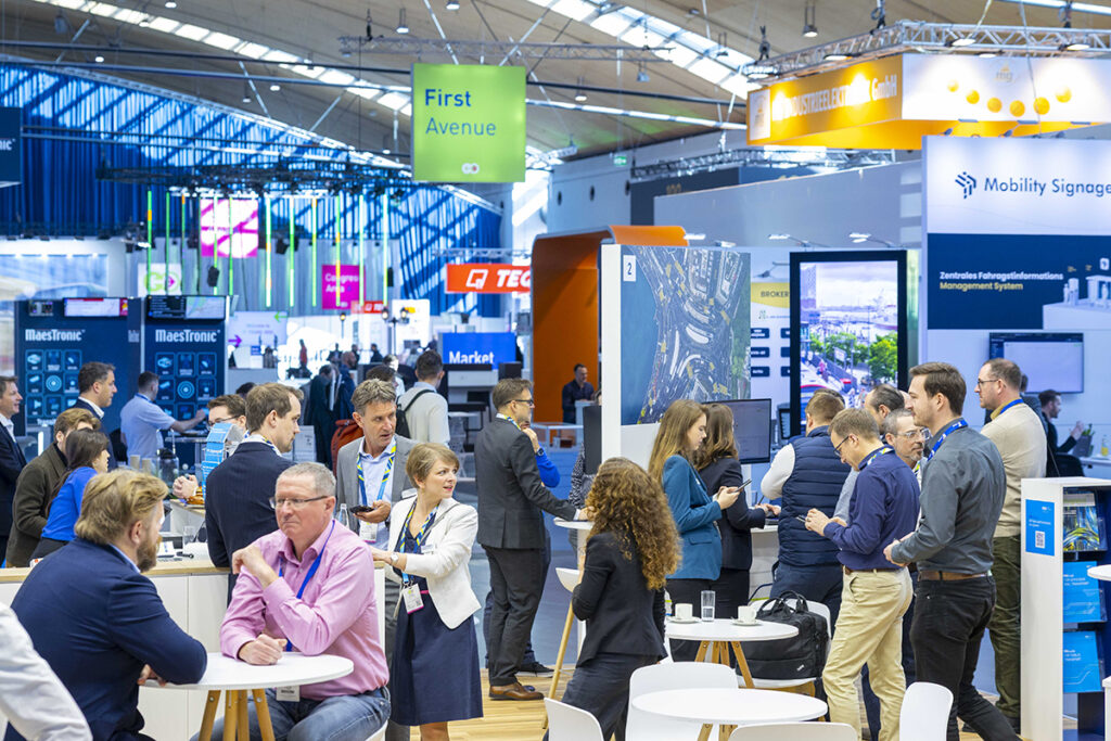 Zahlreiche Personen sitzen und stehen in einer Messehalle.