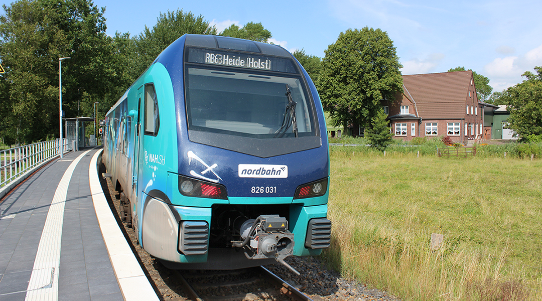 Ein Zug hält vor einer Wiese und einem Haus an einem Bahnsteig.