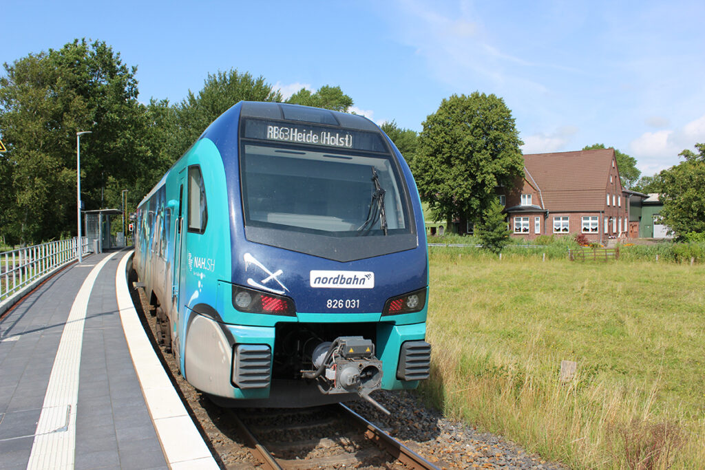 Ein Zug hält vor einer Wiese und einem Haus an einem Bahnsteig.
