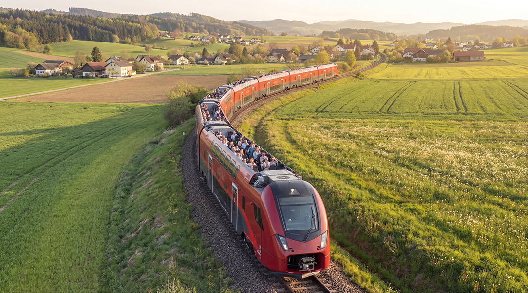 Ein Zug mit offenem Dach fährt durch Wiesen und an Häusern vorbei.