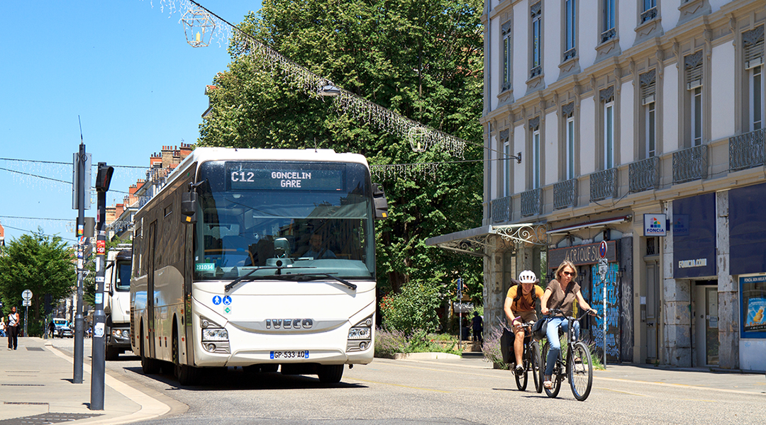 Ein Bus und Radfahrer fahren vor einem Haus und Bäumen auf einer Straße.
