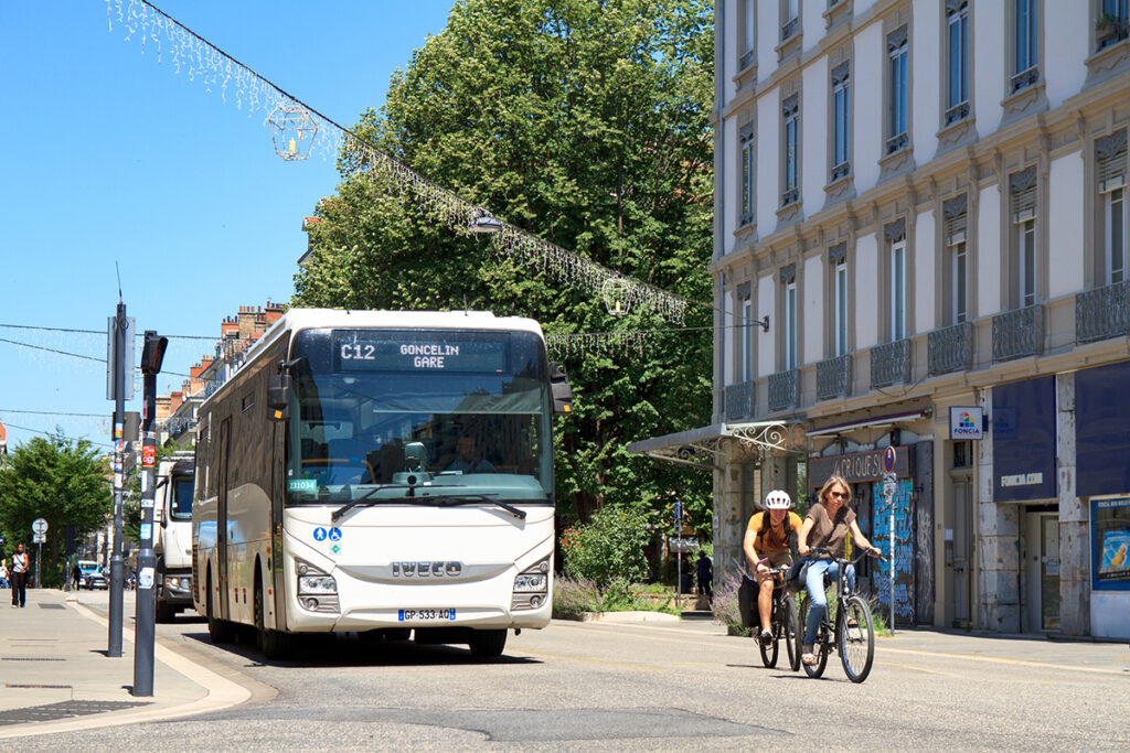 Ein Bus und Radfahrer fahren vor einem Haus und Bäumen auf einer Straße.