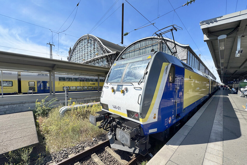 Ein Zug mit Doppelstockwagen steht vor einer Bahnhofshalle an einem Bahnsteig.