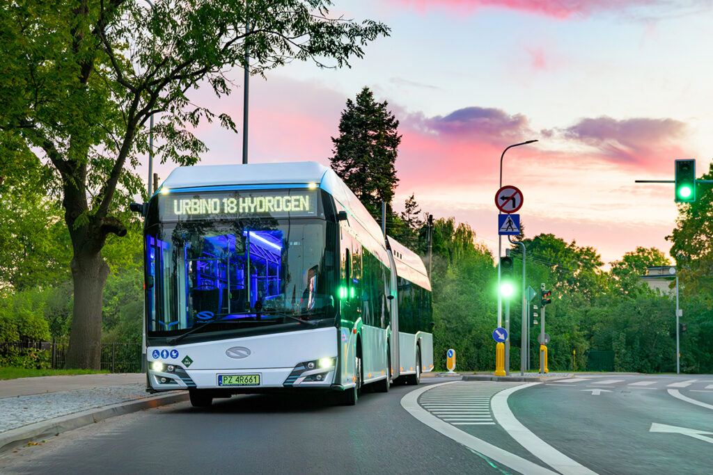 Ein Wasserstoff-Gelenkbus fährt vor Bäumen auf einer Straße.