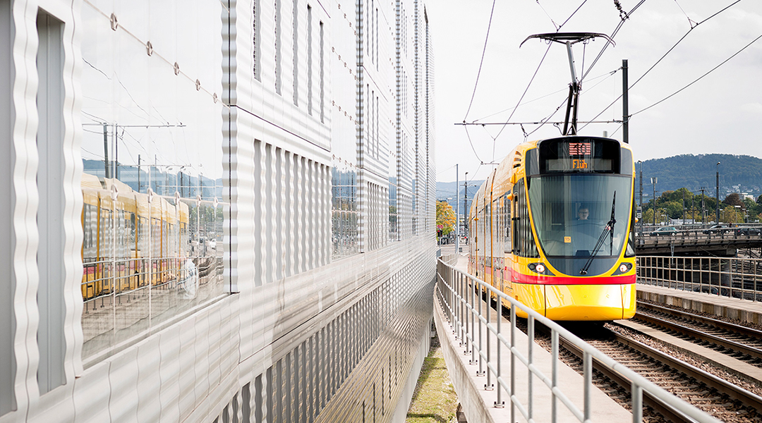 Eine Stadtbahn fährt auf einer Brücke an einem Hochbaus entlang.