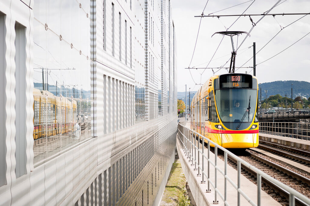 Eine Stadtbahn fährt auf einer Brücke an einem Hochbaus entlang.