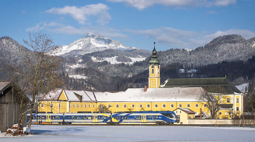 Ein Zug fährt vor einer Kirche und vor Bergen entlang. Es liegt Schnee.