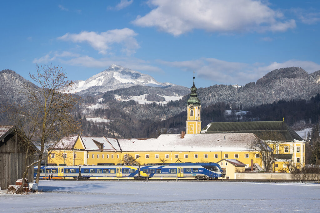 Ein Zug fährt vor einer Kirche und vor Bergen entlang. Es liegt Schnee.