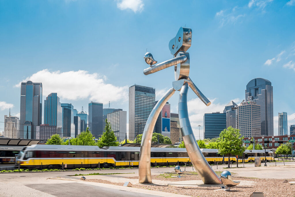 Eine übergroße Statue steht vor einer Stadtkulisse mit einer Straßenbahn im Vordergrund.