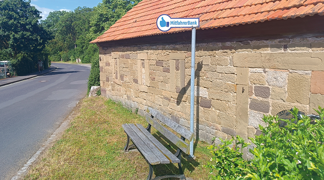 Eine Bank mit Haltestellenschild steht vor einer Hauswand an einer Straße.