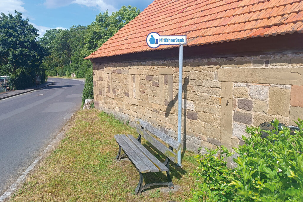 Eine Bank mit Haltestellenschild steht vor einer Hauswand an einer Straße.