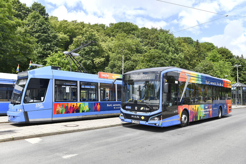 Eine Straßenbahn und ein Bus, beide in Regenbogenfarben beklebt, stehen nebeneinander. 