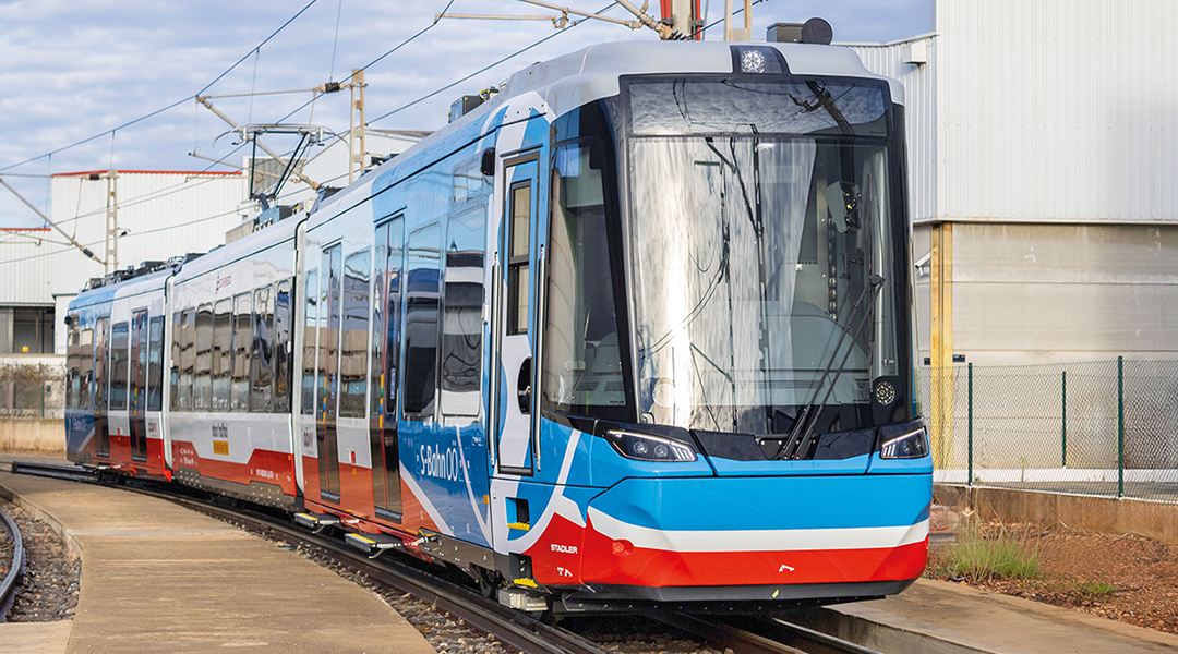 Eine Zweisystem-Straßenbahn steht vor einer Halle auf einem Gleis.