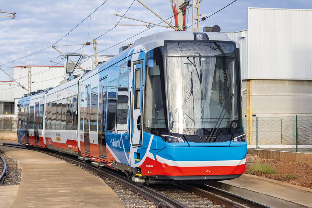 Eine Zweisystem-Straßenbahn steht vor einer Halle auf einem Gleis.
