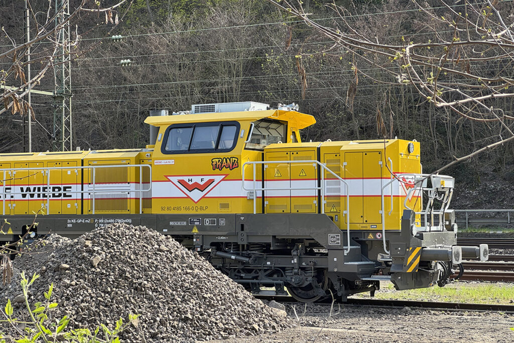 Ein Diesellok steht hinter einem Schotterhaufen vor Bäumen.