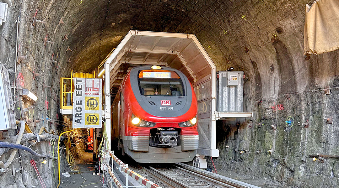 Ein Zug fährt unter einer Baumaschine durch einen Tunnel.