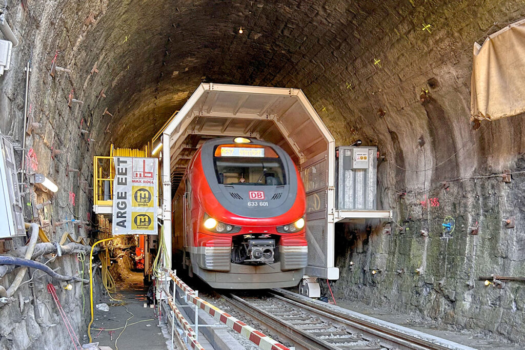 Ein Zug fährt unter einer Baumaschine durch einen Tunnel.