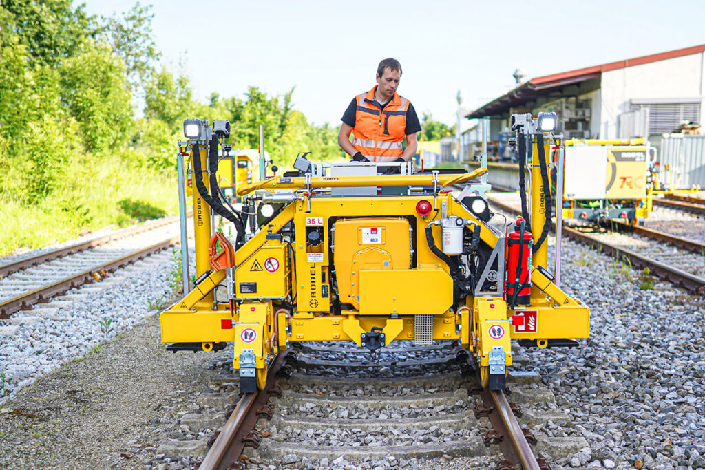 Eine Person steht auf einer kleinen Gleisbaumaschine auf einem Gleis.