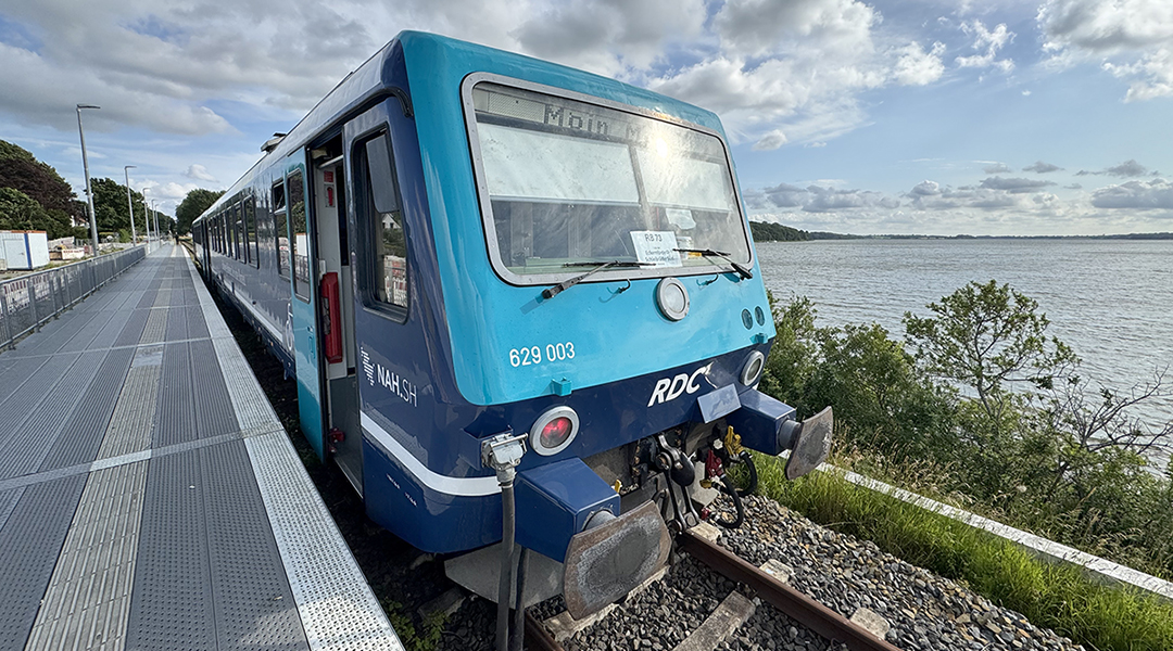 Ein Zug steht an einem Bahnsteig, dahinter befindet sich ein Gewässer.