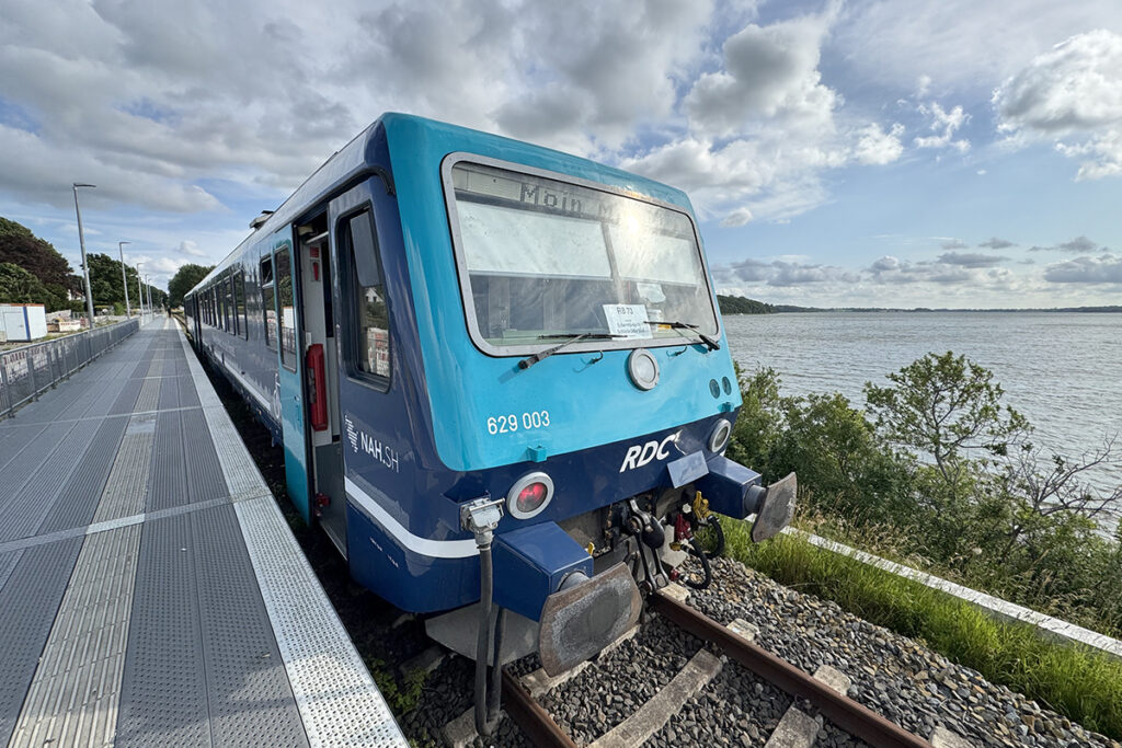 Ein Zug steht an einem Bahnsteig, dahinter befindet sich ein Gewässer.