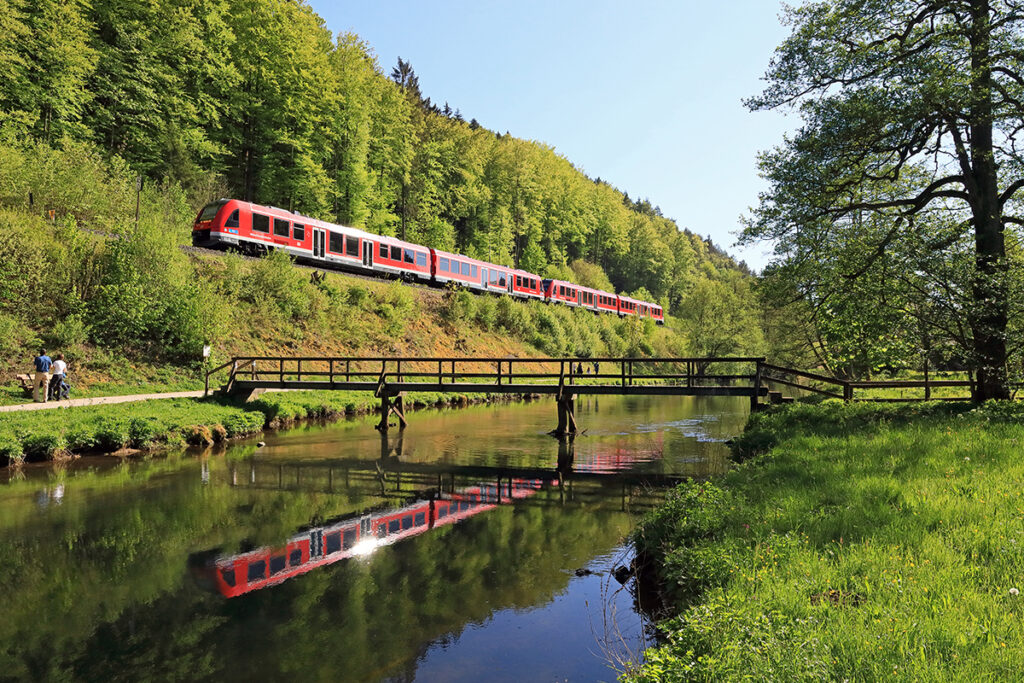 Ein Zug fährt an einem Fluss entlang und spiegelt sich im Wasser.