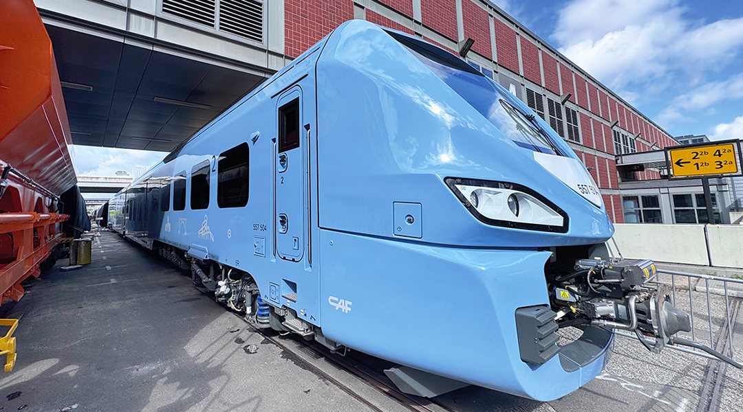 Ein Batteriezug steht neben einem Güterwagen vor einer Messehalle.