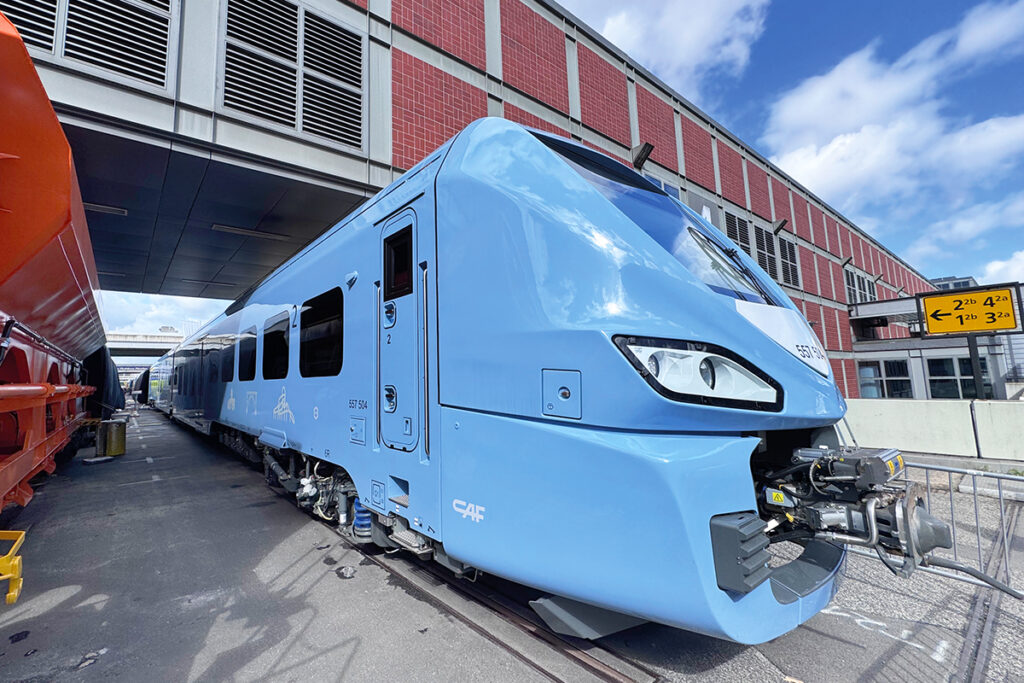 Ein Batteriezug steht neben einem Güterwagen vor einer Messehalle.