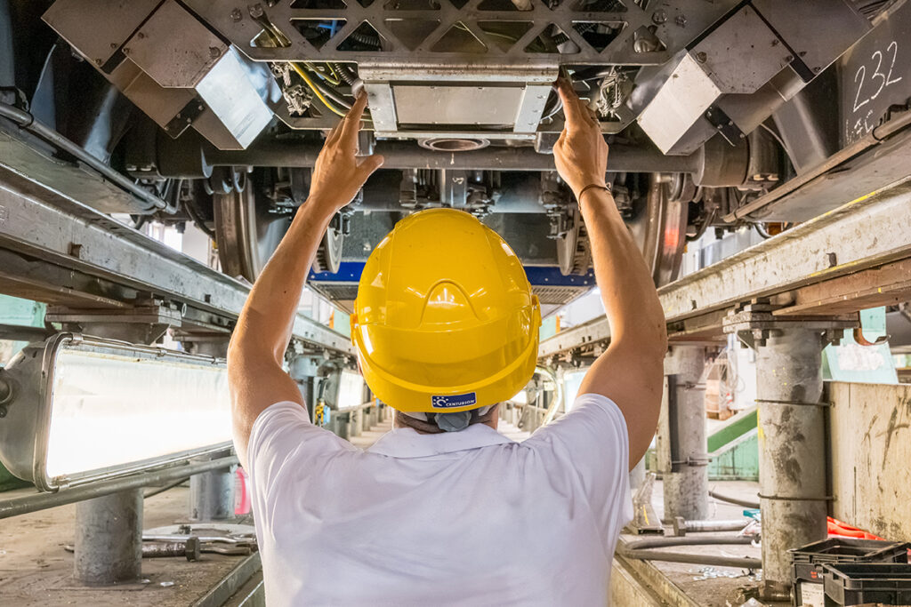 Eine Person steht unter einem Eisenbahnfahrzeug in einer Grube und arbeitet daran. 