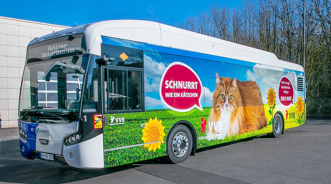 Ein Elektrobus mit einer Katze auf der Seite steht vor einer Halle.
