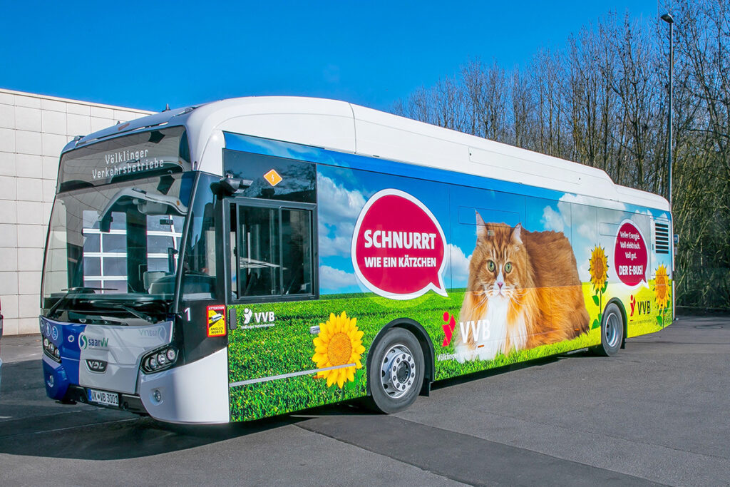Ein Elektrobus mit einer Katze auf der Seite steht vor einer Halle.