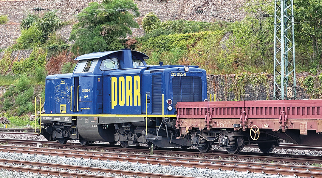 Eine Diesellok steht mit einem Wagen auf einem Gleis vor einer Mauer.