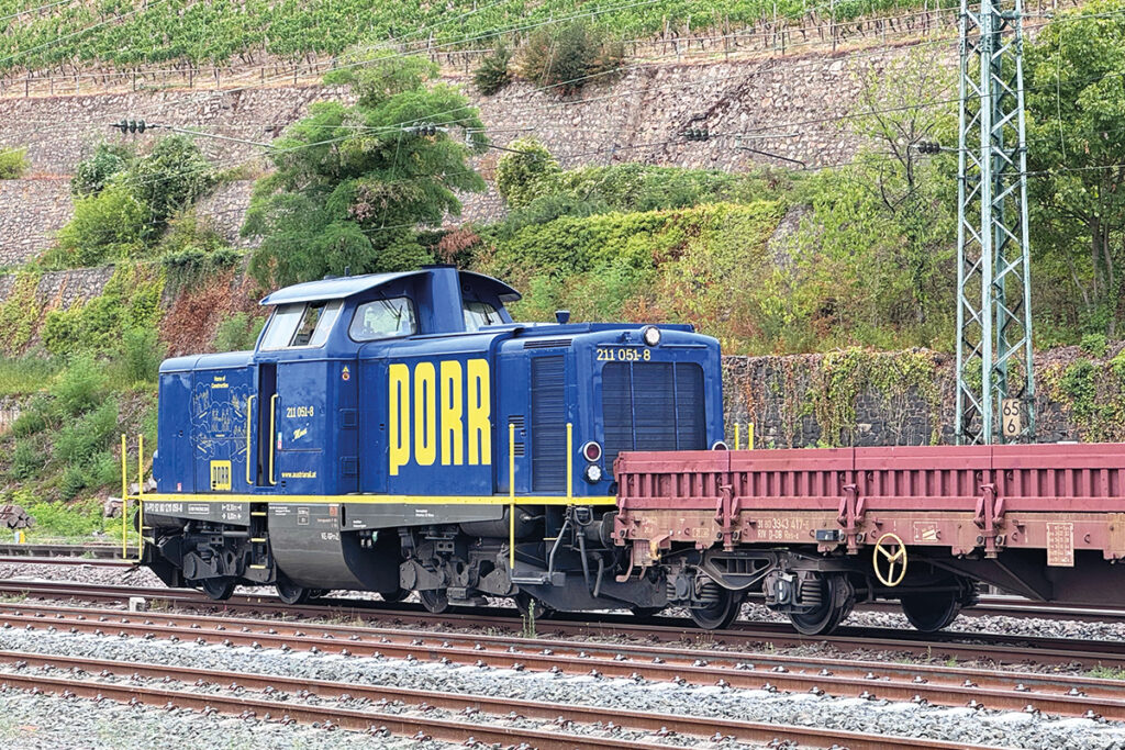 Eine Diesellok steht mit einem Wagen auf einem Gleis vor einer Mauer.