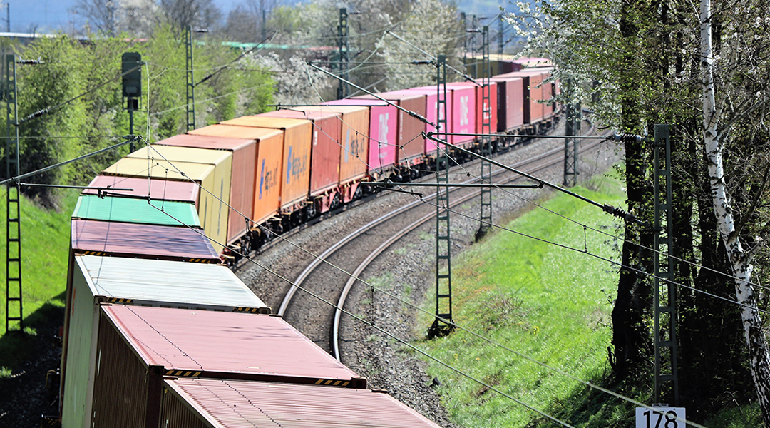Ein Containerzug fährt auf einer zweigleisigen, kurvenreichen Strecke an Bäumen entlang.