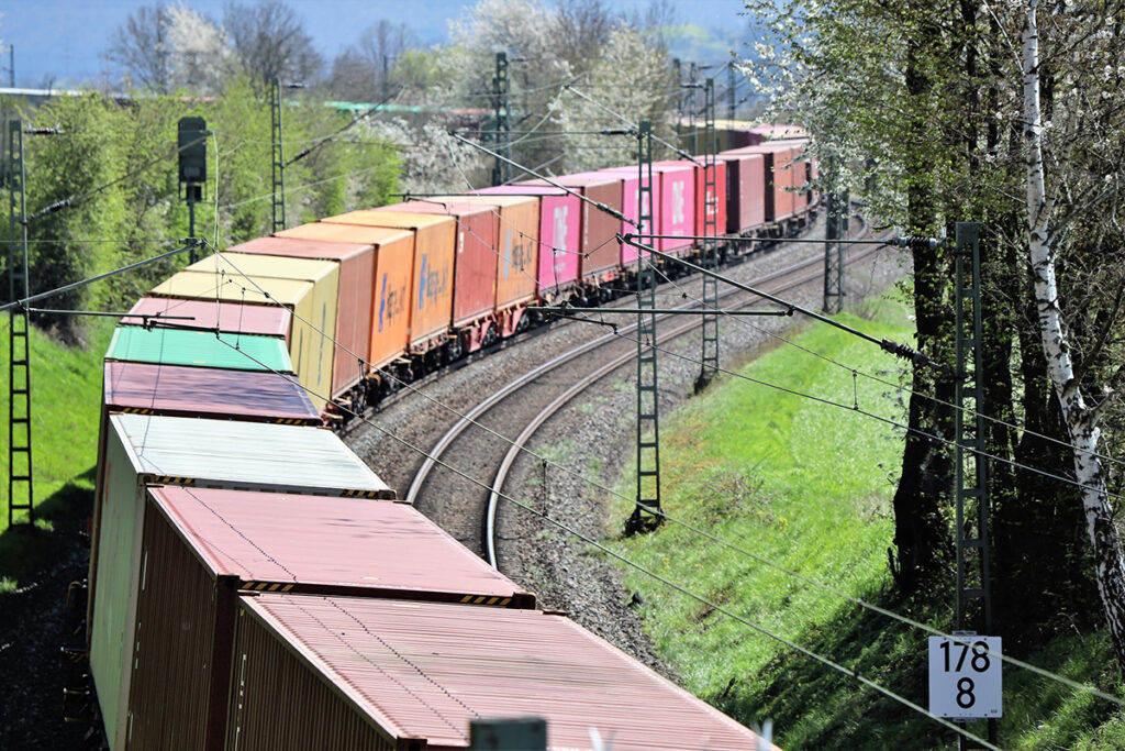 Ein Containerzug fährt auf einer zweigleisigen, kurvenreichen Strecke an Bäumen entlang.